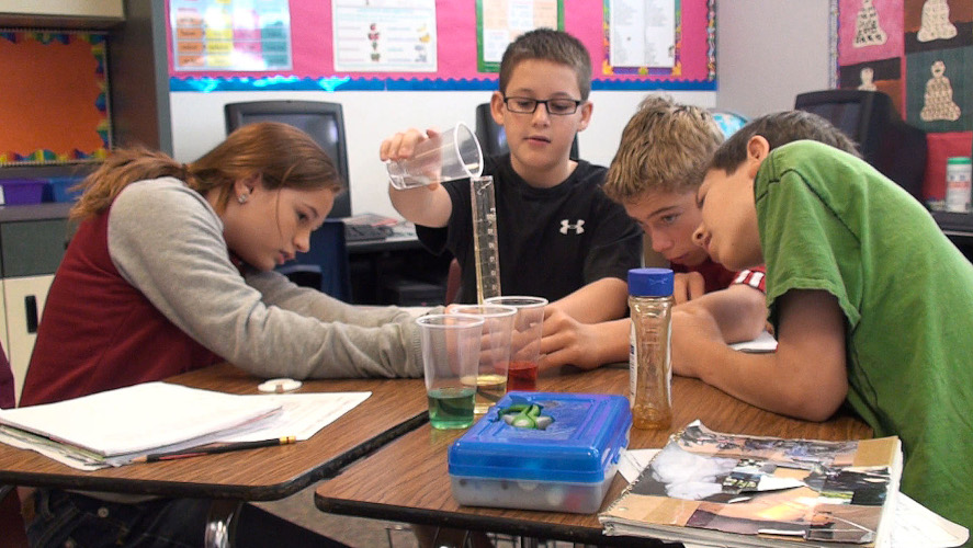 En elev häller vätska i mätglas medan tre elever observerar. 