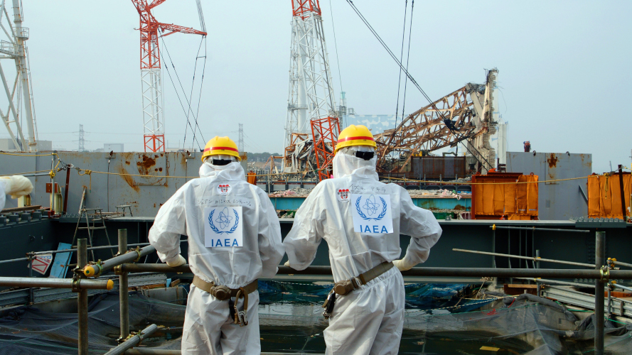 Två personer i skyddsdräkter med IAEA på ryggen står framför kärnkraftverket i Fukushima.