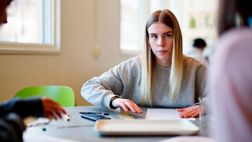Flicka sitter vid skrivbord med två personer i förgrunden.