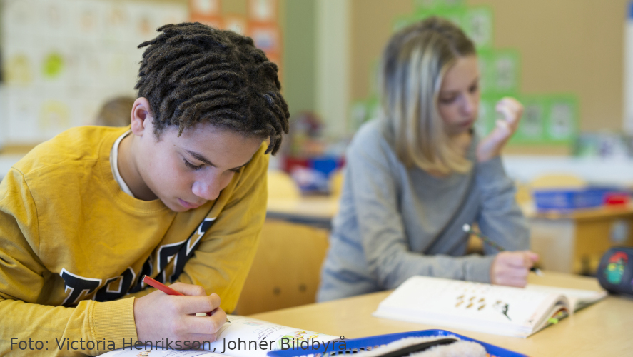 Pojke i förgrund och flicka i bakgrund arbetar med penna och arbetshäfte.